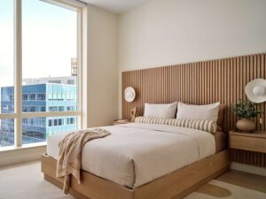 Modern bedroom interior design with wooden bed, wooden panel headboard wall, side tables, and large window with city view.